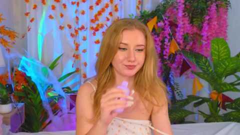 Video of a young Caucasian woman with long blonde hair, wearing a floral-patterned dress, holding a lighter, surrounded by vibrant tropical plants and colorful decorations in a brightly lit room.
