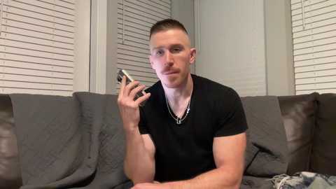Video of a young, fair-skinned, muscular man with short hair and a mustache, wearing a black shirt, sitting on a gray couch, holding a phone, with white blinds in the background.