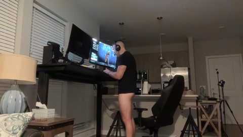 Video of a man in a black t-shirt and no pants, sitting at a desk with a gaming setup, surrounded by a modern kitchen and living space.