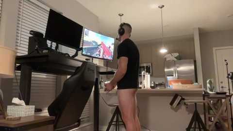 A video of a muscular, shirtless man in black shorts, wearing headphones, playing a video game on a TV console in a modern, dimly-lit living room.