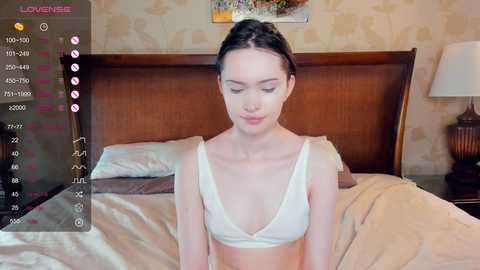 Media: A video of a young, slim woman with fair skin and dark hair in a bun, wearing a white bra, lying on a bed in a cozy, vintage bedroom.