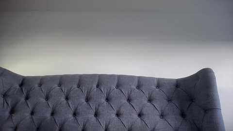 A video of a plush, gray velvet tufted headboard with a curved design against a plain, light gray wall. The headboard features a diamond pattern and appears soft to the touch.