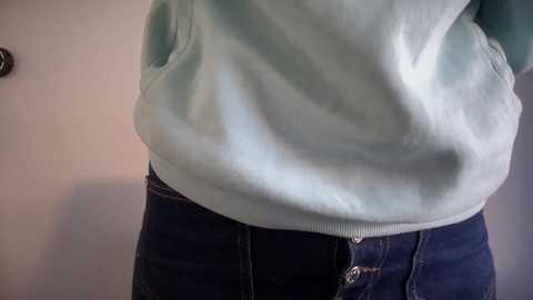 Video of a person wearing a light blue hoodie and dark blue jeans, hands in pockets. Background is a plain, light-colored wall.