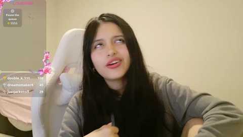 Video of a young woman with long black hair, fair skin, and pink lips, sitting on a white gaming chair with pink flowers, wearing a grey shirt, in a simple, beige-walled room.