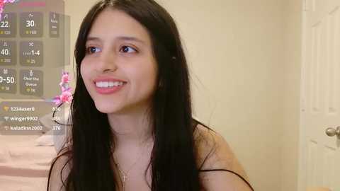 Video of a smiling Latina woman with long black hair, wearing a sleeveless top, standing in a bedroom with a pink floral curtain and a white door.