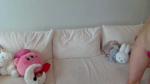 Video of a plush pink Kirby and white rabbit toys on a beige couch. A nude person with a tattooed arm is partially visible in the foreground.