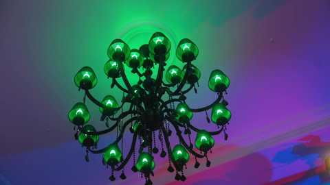 Media: Video of an ornate black chandelier with 16 green glass globes, emitting soft light, hanging in a room with a colorful, abstract background featuring purple, pink, and blue hues.