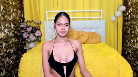 Video of a young woman with medium skin tone and long black hair, wearing a black halterneck top, sitting on a bed with yellow sheets and pillows, surrounded by gold-patterned curtains.