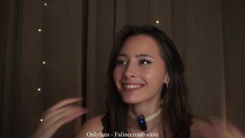 Video of a smiling woman with long brown hair, wearing a black dress with a blue necklace, against a wooden backdrop with string lights.