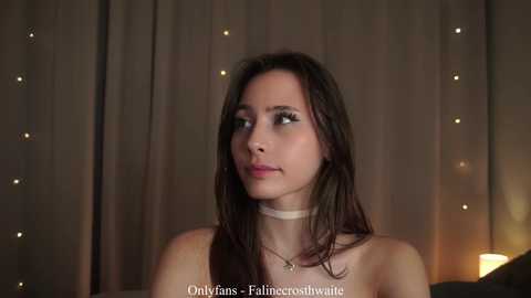 Video of a young woman with long dark hair and light skin, wearing a choker necklace, looking contemplative in a dimly lit room with fairy lights.