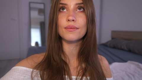 Video of a young woman with fair skin, long brown hair, and green eyes, wearing a white off-shoulder top, standing in a modern bedroom with a gray bed and white walls.