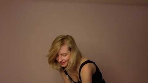 Video of a young, fair-skinned woman with shoulder-length blonde hair, wearing a black tank top, laughing joyfully with eyes closed, set against a plain, beige wall background.