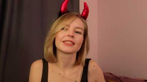 Media: Video of a blonde woman with shoulder-length hair, wearing black horns and a black tank top, smiling, indoors with a blurred background.