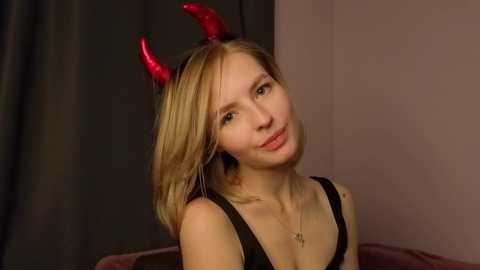 Media: Video of a blonde woman with fair skin, wearing a black tank top, and red devil horns, smiling against a dark curtain background.