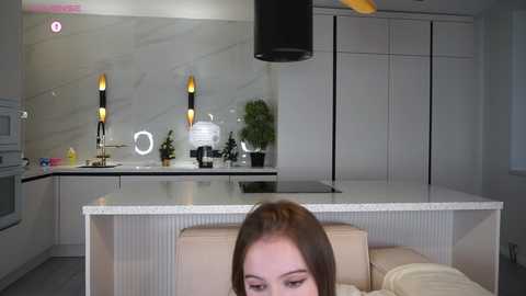 Video of a modern, minimalist kitchen with white cabinets, marble countertops, and a beige couch. A woman with brown hair sits on the couch, facing the camera.