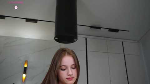 Video of a young Caucasian woman with fair skin and long, straight brown hair, wearing a black top, standing in a modern room with a sleek, minimalist aesthetic.