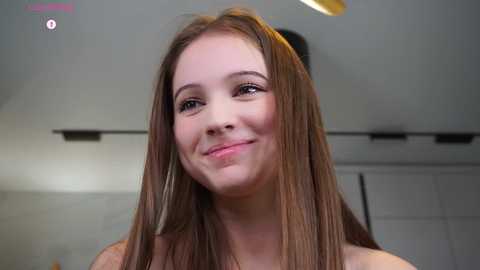 Media: Video of a young Caucasian woman with long brown hair and fair skin, smiling slightly, standing indoors with white walls and a ceiling light fixture in the background.