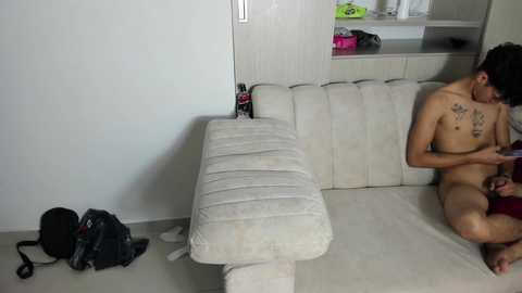 Video of a young Asian man, shirtless, sitting on a beige sofa, reading a book, with a black backpack and a black bag on the floor.