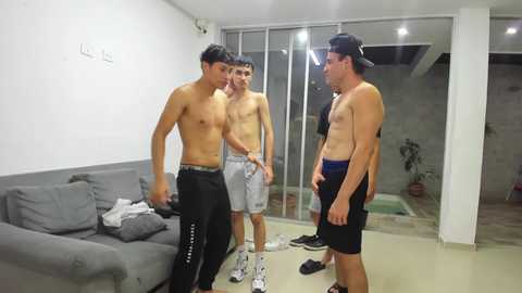 Video of four shirtless young men in a gym; one wears black shorts, two wear grey shorts, and one wears a cap and blue shorts.