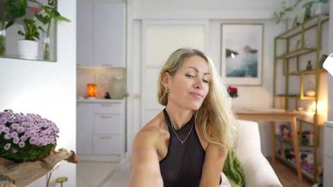 Video of a blonde Caucasian woman with long hair in a black halter top, smiling in a bright, modern kitchen with white cabinets, greenery, and a vase of purple flowers on a wooden table.
