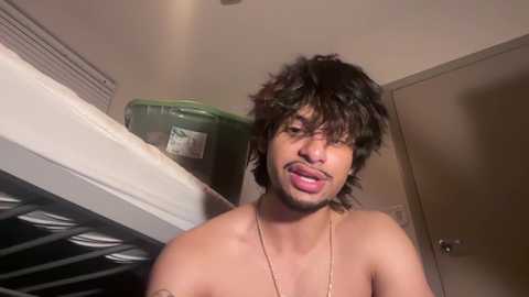 Video of a shirtless man with curly hair, dark skin, and a playful expression licking his lips. Background includes a green trash can, a door, and a shelf.