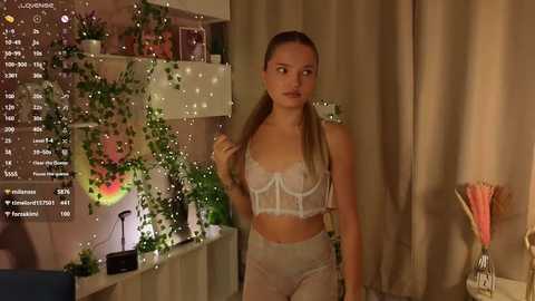 Media: Video of a young woman in white lingerie, standing indoors with green plants, a TV screen displaying social media stats, and a vase of flowers in the background.