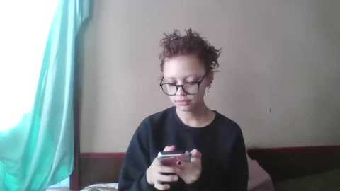 A video of a young person with curly brown hair, wearing glasses, black t-shirt, and headphones, sitting indoors by a window with green curtains, focused on a smartphone.