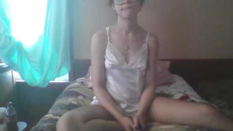 Video of a fair-skinned woman with glasses, wearing a white satin camisole, sitting cross-legged on a bed with a floral quilt in a dimly lit room with green curtains.