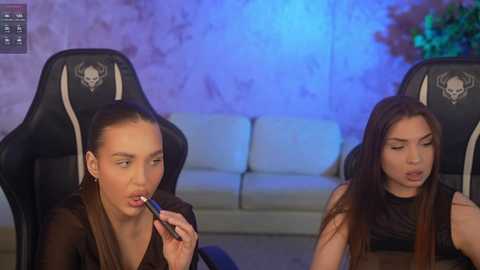 Video of two women, both with long brown hair, sitting in black gaming chairs, applying lipstick in a dimly lit room with blue and purple lighting.