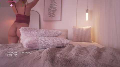 A video of a woman in a maroon bra and thong, standing with her back to the camera, in a cozy bedroom with white walls, a textured blanket, and a hanging light.