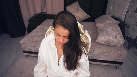Video of a young woman with long, straight black hair, wearing a white bathrobe, sitting on a plush, beige chaise lounge in a modern, softly lit bedroom with gray walls and soft furnishings.