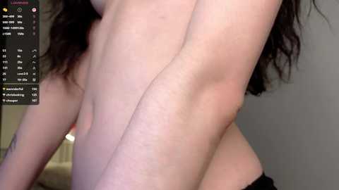 Video of a pale-skinned person with long, wavy brown hair, partially topless, showing an armpit and part of a breast. Background includes a blurred, neutral-colored room with a digital screen displaying weather information on the left.