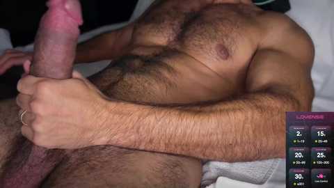 Video of a nude, muscular man with tan skin, holding his erect penis, on a bed with white sheets. Background includes a digital screen displaying \"Loadz\" game statistics.