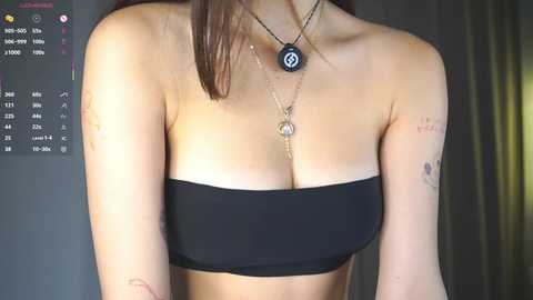 Video of a woman with light skin and brown hair, wearing a black strapless top, a black pendant necklace, and a black beaded bracelet. Tattoos on her arms. Background includes a dark wall with a calendar and a black curtain.