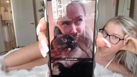 Video of a blonde woman with glasses, lying on a white fur rug, taking a selfie with a black cat, background shows modern interior with bookshelves.