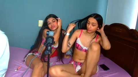 Video of two Latina women with long black hair, wearing matching pink lingerie, sitting on a bed, combing each other's hair, in a dimly lit room.