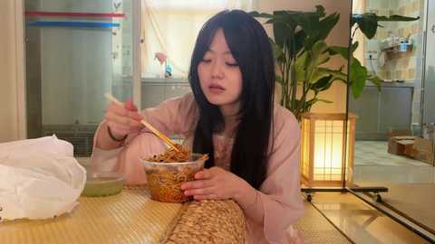 A video of an Asian woman with long black hair, wearing a pink kimono, eating ramen noodles from a bowl with chopsticks, seated at a wooden table in a cozy, traditional Japanese room with bamboo and paper lanterns.