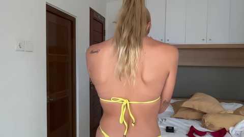 Video of a blonde woman with a sun-kissed tan, wearing a yellow bikini, standing in a bedroom with white cabinets and a messy bed.