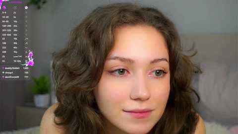 Video of a young Caucasian woman with wavy brown hair and fair skin, wearing minimal makeup, looking directly at the camera. Background shows a gray wall with a potted plant and a digital clock display.