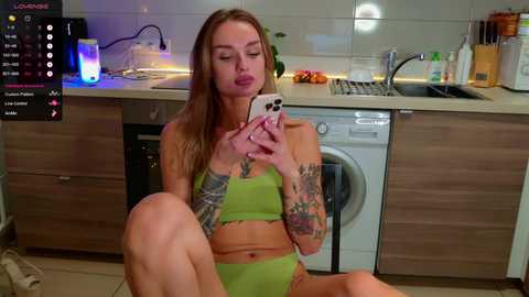 Media: A young woman with light skin and long, blonde hair sits in a modern kitchen, wearing green lingerie. She has tattoos on her arms and is holding a smartphone. The kitchen is well-lit with a stainless-steel sink and cabinets.