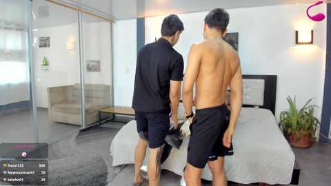 Video of two shirtless men in black shorts, one holding a dog, in a modern, minimalistic bedroom with a bed, potted plant, and framed artwork on white walls.