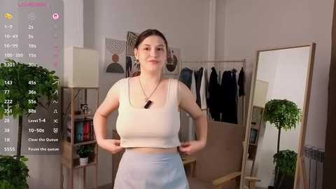 Video of a smiling, fair-skinned woman with medium-length brown hair, wearing a white crop top and light blue skirt, standing in a modern, minimalistic room with a mirror and plants.