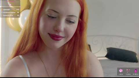 Media: Video of a young, fair-skinned woman with long, vibrant orange hair, wearing a white tank top, smiling slightly. Background shows a white bed with a black pillow.
