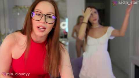 Video of a woman with long, straight, light brown hair and glasses wearing a red halter dress, laughing. In the background, a woman in a white dress dances, and another woman in a white dress looks confused.