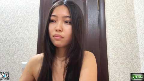 Media: Video of an Asian woman with long, straight black hair, wearing a black top, against a textured white and beige wallpaper. She has a neutral expression.