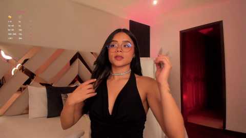Video of a confident woman with medium skin tone, long black hair, and glasses, wearing a black halter dress and choker, posing in a modern, dimly-lit room with geometric patterns and red lighting.