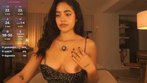 A video of a young woman with long, wavy black hair, wearing a black, polka-dotted, strapless top that exposes her breasts, sitting in a dimly lit living room.