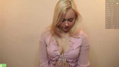 Video of a blonde Caucasian woman with straight hair, wearing a light pink blouse with a low neckline, revealing cleavage. She has a soft expression and is standing against a beige wall.