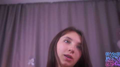 Video of a young woman with long brown hair, light skin, and blue eyes, looking upward with a neutral expression. She stands in front of gray curtains in a dimly lit room.