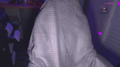 Video of a person sitting on a textured gray blanket in a dimly lit room, with a purple light casting shadows. Background shows a messy kitchen counter and a closed door.